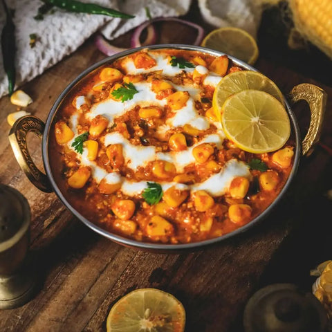 A bowl of Indian chickpea curry, garnished with coriander, cream, and lemon slices, is enhanced with Gustus Vitaes Better Than Butter Seasoning Set: 2 Pack. Sliced onions, lemon, and green chili are in the background on a wooden table.