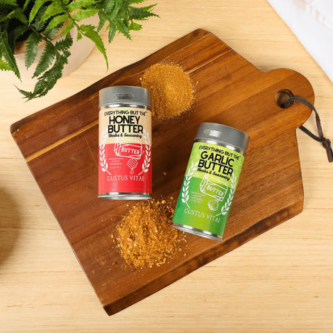 The Gustus Vitae Better Than Butter Seasoning Set: 2 Pack features Honey Butter and Garlic Butter tins on a wooden board with spices, a plant, and a white cloth atop a light wood surface.