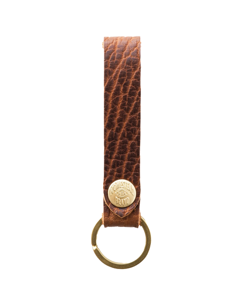 The Bison Standard Key Fob No. 1603 by Homestead Brand features rugged, textured brown full-grain leather, a gold metal ring, and a gold-engraved button—an ideal American-made keychain for everyday use.