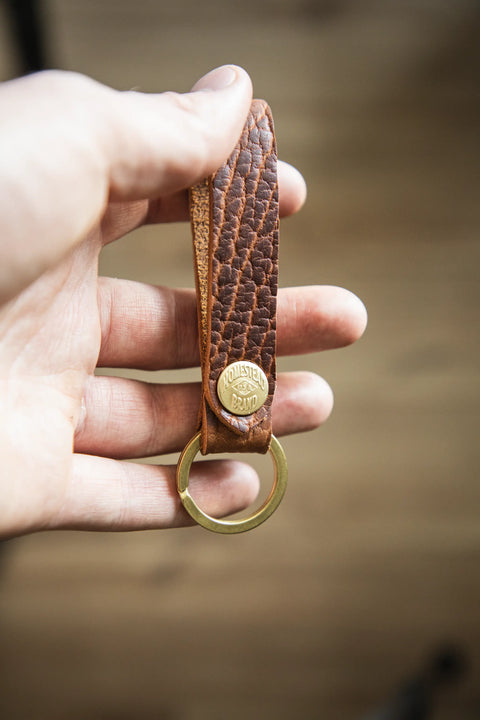 A hand holds the Bison Standard Key Fob No. 1603 by Homestead Brand, crafted from brown full-grain leather with a textured finish, a brass keyring, and a round brass button engraved with the brand. The background is softly blurred.