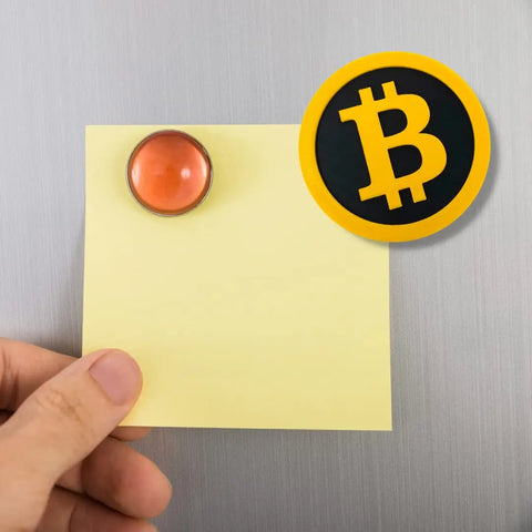 A hand holds a yellow sticky note on a metallic surface, secured by a red magnet. Above it is the Chatelet Manufacturing Bitcoin Fridge Magnet—a decorative magnet for cryptocurrency fans that adds unique style to your space.