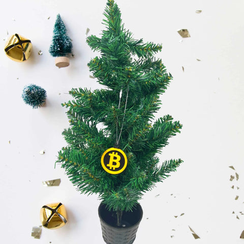 A black-potted mini Christmas tree is topped with the Chatelet Manufacturing Bitcoin Christmas Ornament—perfect for cryptocurrency fans. Gold jingle bells, green trees, and gold confetti accent the festive white display. Made in the USA.