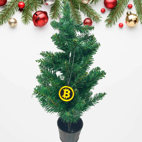 A small potted Christmas tree features a Chatelet Manufacturing Bitcoin Christmas Ornament—perfect for cryptocurrency fans. Pine branches with red, gold, and white ornaments hang above on a white background. Made in the USA.