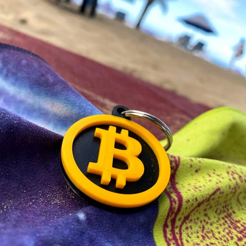 The Bitcoin Keychain by Chatelet Manufacturing, a decorative accessory for cryptocurrency fans, is shown in yellow and black on a beach towel with sand, umbrellas, and people blurred in the background.