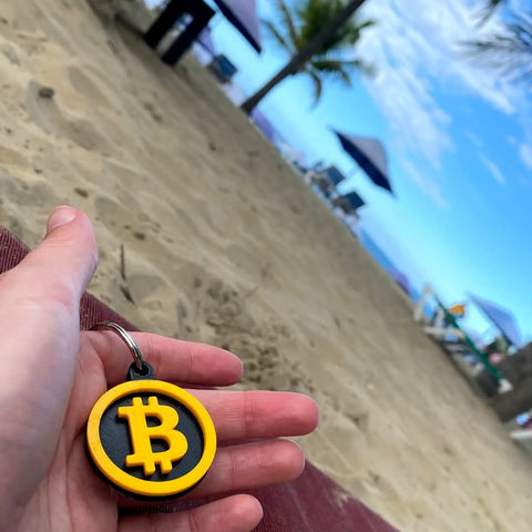 A hand holds the Chatelet Manufacturing Bitcoin Keychain, featuring a yellow symbol, on a sandy beach with palm trees, umbrellas, and lounge chairs—an ideal decorative accessory for cryptocurrency fans.