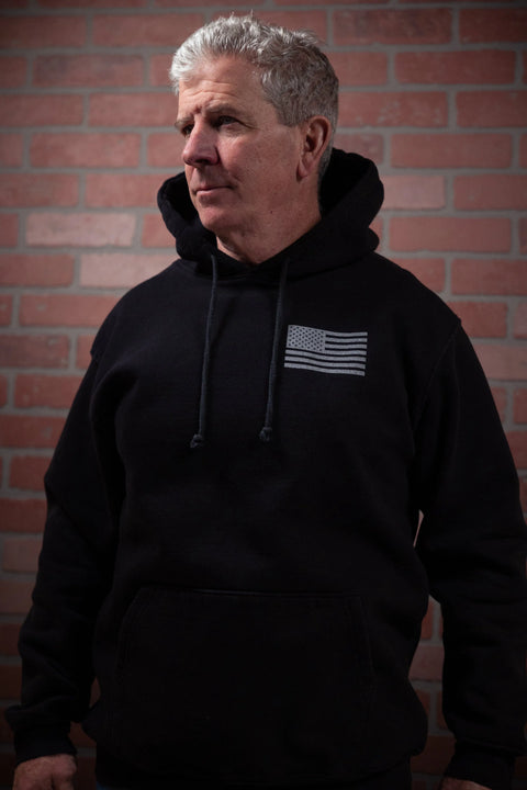 An older man with gray hair wears the Proudly USA American Flag Pullover Hoodie Sweatshirt (Black), standing before a red brick wall and gazing to the side.