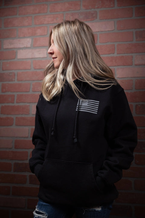 A woman with long blonde hair stands in front of a red brick wall, wearing the Proudly USA American Flag Pullover Hoodie Sweatshirt (Black) with distressed blue jeans, looking off to the side with her hands in her pockets.
