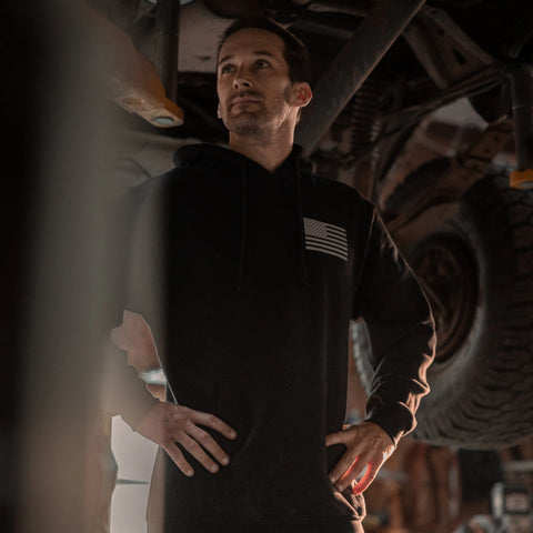A man wearing the Proudly USA American Flag Pullover Hoodie Sweatshirt (Black) stands under a raised vehicle in a garage, looking up with his hands on his hips.
