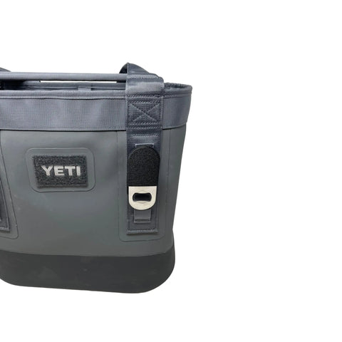 A partial view of a gray YETI cooler bag with black trim displays the Chatelet Manufacturing Black Bottle Opener Attachment, compatible with soft YETI coolers and backpacks, secured to the front as a convenient removable accessory.