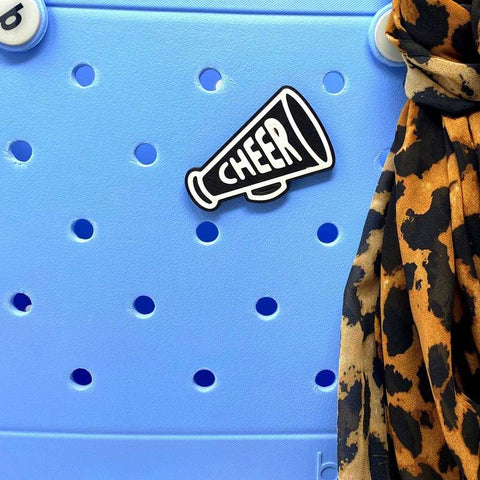 The BOGLETS - Black Megaphone Charm by Chatelet Manufacturing, featuring CHEER in white, is shown on a light blue Bogg Bag with a brown and black leopard print scarf—great accessories for cheer teams.