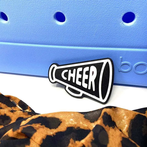 The BOGLETS - Black Megaphone Charm by Chatelet Manufacturing, featuring CHEER in white, is attached to a blue Bogg Bag with leopard print fabric peeking below—a fun accessory for cheer teams.