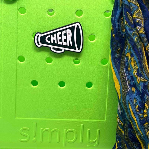A green surface with simply at the bottom, a Chatelet Manufacturing BOGLETS black megaphone charm for Bogg Bags reading CHEER, and blue-yellow patterned fabric on the right—a fun pick for cheer teams.