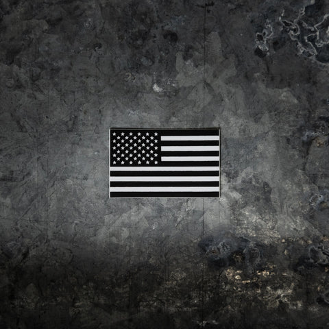 The Proudly USA American Flag Sticker (Black and White, 2.75 x 1.71) stands out against a dark, textured concrete wall, creating a striking patriotic focal point with its bold design.