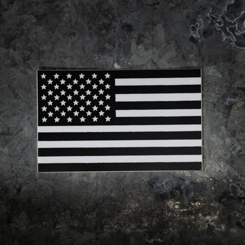 The Proudly USA American Flag Window Decal (Black and White, 5.5 x 3.42) stands out on a dark, textured concrete surface—ideal for adding patriotic flair to your car or truck window.