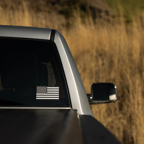 A silver pickup, parked outdoors by tall dry grass, displays a Proudly USA American Flag Window Decal (Black and White, 5.5 x 3.42) on its rear window.