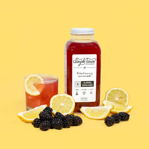 A bottle of Simple Times Mixers Blackberry Lemonade sits beside a glass of lemonade cocktail with a lemon slice, surrounded by blackberries and lemon wedges on a solid yellow background.