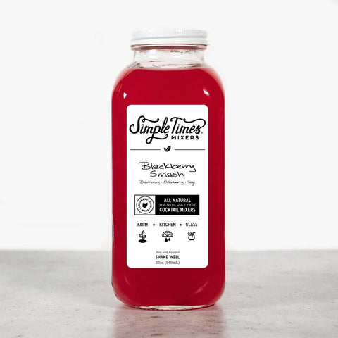 A clear 32oz bottle of Simple Times Mixers Blackberry Smash is filled with red non-alcoholic cocktail mix and sits on a light surface. The label highlights all-natural blackberry, elderberry, and sage flavors.