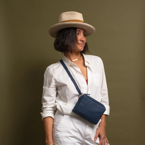 A woman stands against an olive green background wearing a beige hat, white button-up shirt, and white pants, styled with the CASUPO Fanny Pack - Blue worn crossbody.