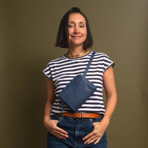 A woman wearing a striped t-shirt, blue jeans, and a brown belt smiles at the camera. She carries the CASUPO Fanny Pack - Blue as a crossbody and stands against an olive-green background.