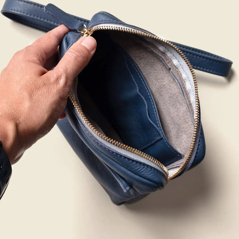 A hand holds open the CASUPO Fanny Pack - Blue, made from sustainable leather with a gold zipper, showing its empty, light-colored fabric interior.