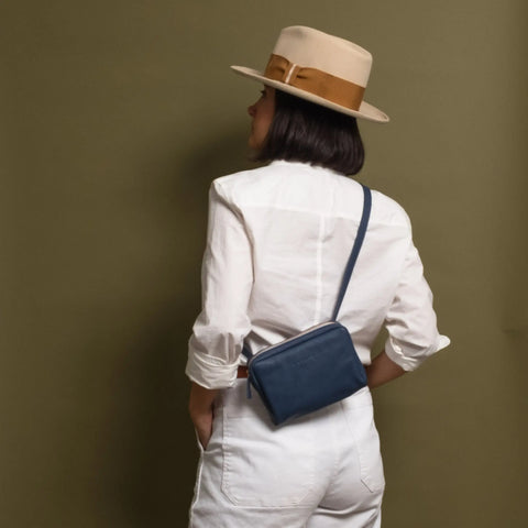 A person stands with their back to the camera, wearing a beige hat, white long-sleeve shirt, and white pants. Draped over their shoulder is the CASUPO Fanny Pack - Blue, set against an olive green background.
