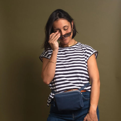 A woman in a black and white striped shirt, blue jeans, and the CASUPO Fanny Pack - Blue smiles while adjusting her sunglasses against an olive green background.
