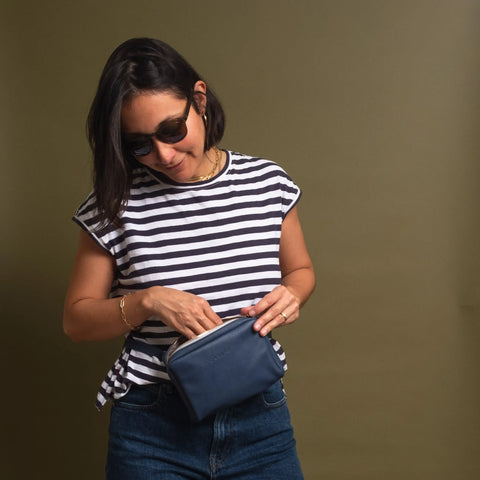 A woman in sunglasses and a black-and-white striped shirt stands against an olive background, looking down as she reaches into the CASUPO Fanny Pack - Blue worn around her waist.