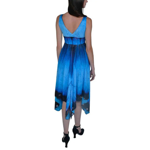 A woman in black heels models the Blue Morpho Butterfly Dress by Spiritex | Sustainable Apparel, featuring a sleeveless design, V-back, and handkerchief hemline, as she stands with her back to the camera against a white background.