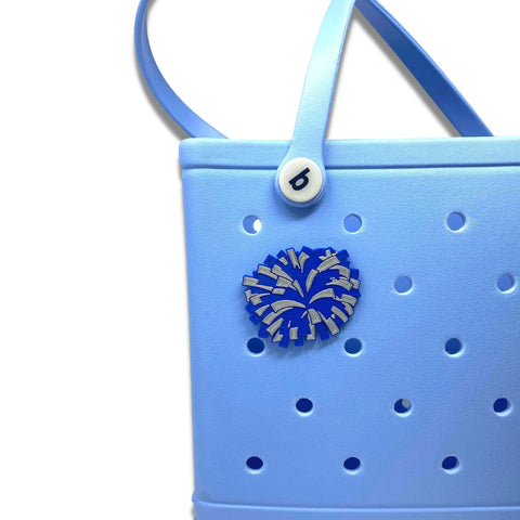 A close-up of a light blue Bogg Bag tote featuring the Chatelet Manufacturing BOGLETS Cheerleading Charm (Blue Pom Pom), a decorative charm compatible with Bogg Bags, and a white button with a lowercase “b” on the handle.