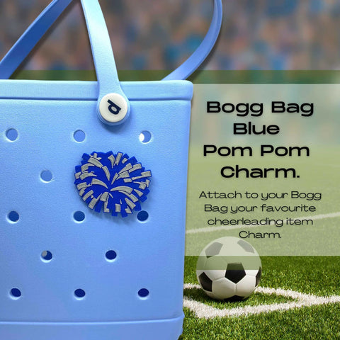 A blue Bogg Bag features the Chatelet Manufacturing BOGLETS Cheerleading Charm (Blue Pom Pom), set on a blurred soccer field with a ball nearby. Text: Add team spirit to your tote with this decorative charm compatible with Bogg Bags!.