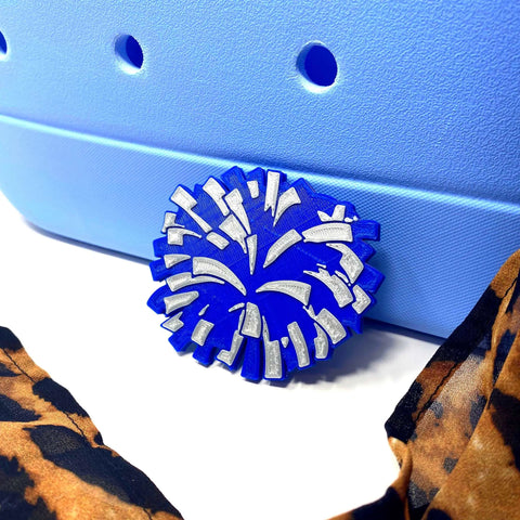 A Chatelet Manufacturing BOGLETS Cheerleading Charm (Blue Pom Pom) is attached to a light blue Bogg Bag with holes, next to brown and black animal print fabric.