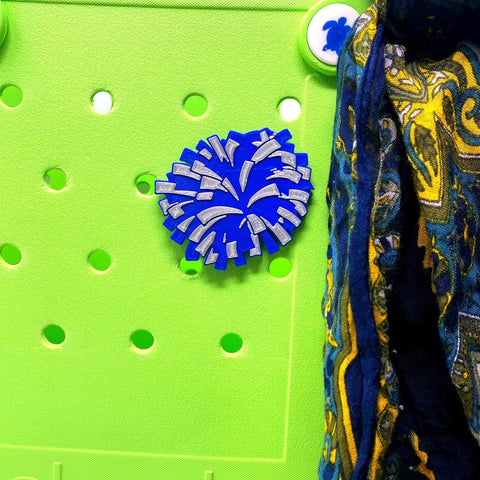 The BOGLETS Cheerleading Charm by Chatelet Manufacturing, featuring a blue pom-pom pin, is shown on a green board with part of a blue and yellow scarf—ideal for accessorizing Bogg Bags and other tote bags.