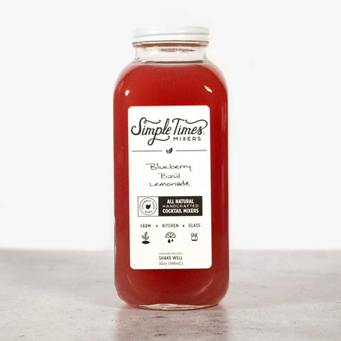 A clear bottle of Simple Times Mixers Blueberry Basil Lemonade, filled with red liquid, sits on a light gray surface. Its white label highlights this non-alcoholic mixer made with all natural ingredients.