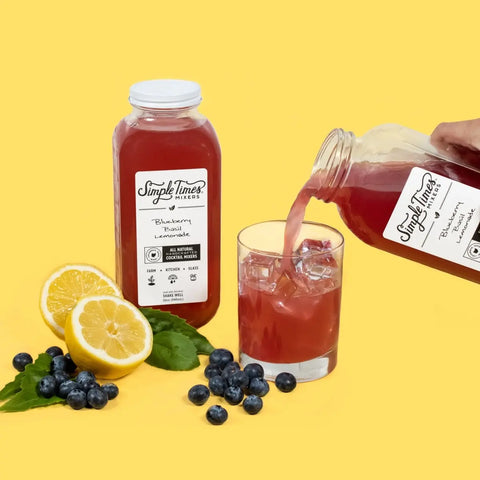 A bottle of Simple Times Mixers Blueberry Basil Lemonade sits beside an ice-filled glass as lemonade is poured in. Lemons, blueberries, and basil garnish the yellow background, highlighting this all-natural non-alcoholic mixer.