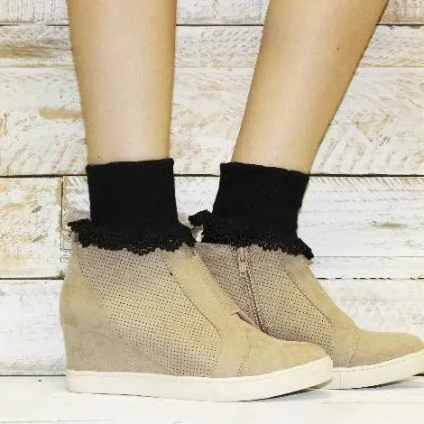 A person wears Catherine Coles BOBBY Lace Cuff Ankle Socks in black with beige wedge sneakers and side zippers, standing before a light wooden background.