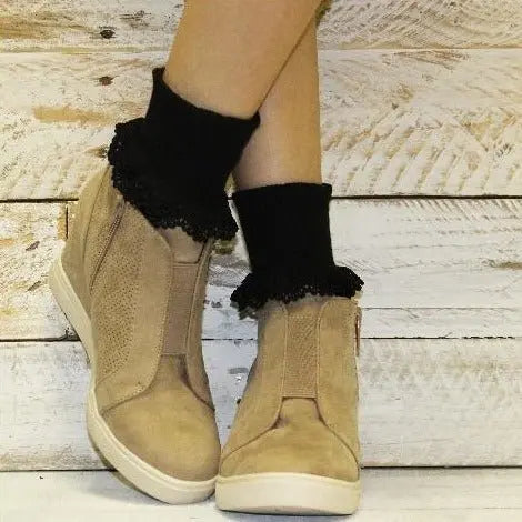 A person stands with legs crossed, wearing beige slip-on high-top sneakers and Catherine Cole BOBBY Lace Cuff Ankle Socks in black, against a rustic white wooden background.