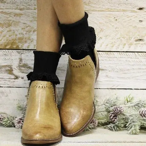 A person stands on a white wooden floor with frosted pine branches, wearing tan leather ankle boots and Catherine Coles BOBBY Lace Cuff Ankle Socks in black.