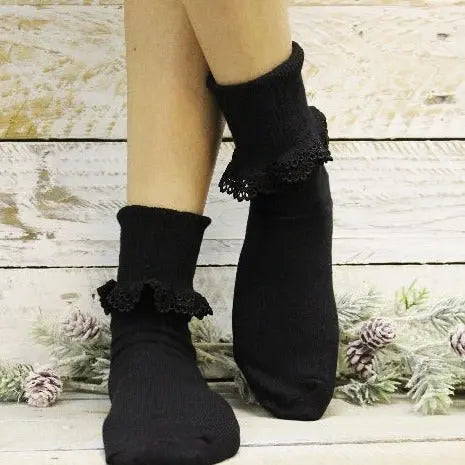 A person models Catherine Cole’s BOBBY Lace Cuff Ankle Socks in black, standing with crossed feet before a white wooden backdrop adorned with frosted pine branches and pinecones.