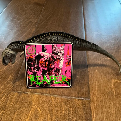 An American Lictor bobcat sticker labeled Reaper with neon pink and green accents rests on a wooden floor, held by a toy dinosaur.