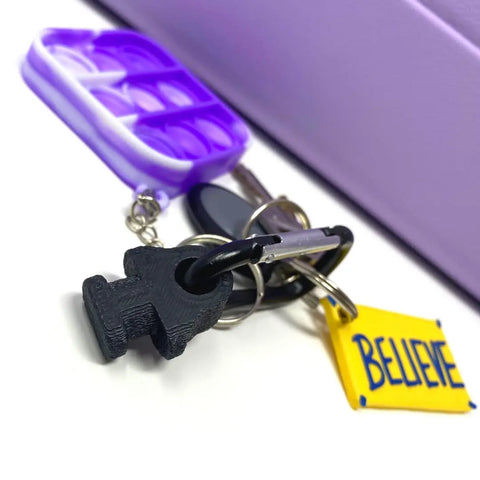 The BOGLETS by Chatelet Manufacturing is a carabiner keys holder charm accessory, shown here with three keychains—a purple pop-it toy, a black figure, and a yellow BELIEVE tag—resting on a white surface.