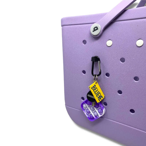 The Chatelet Manufacturing BOGLETS Bogg Bag Carabiner Keys Holder Charm Accessory is a lavender bag with white button accents, a yellow BELIEVE tag, and a purple pop-it fidget toy—perfect for securing keys in your Bogg Bag.