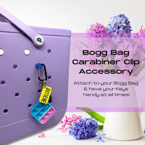 A lavender Bogg Bag holds a colorful pop-it keychain and yellow “BENIE” tag, secured with the Chatelet Manufacturing BOGLETS Carabiner Keys Holder Charm Accessory—keeping valuables organized by pink and purple flowers.