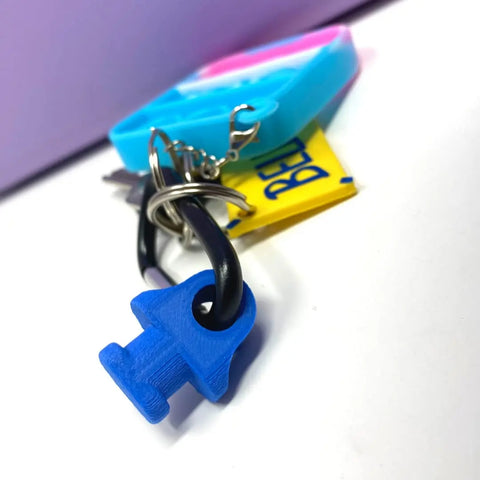 A close-up of the BOGLETS by Chatelet Manufacturing, a carabiner key holder charm accessory for Bogg Bags, featuring a blue gender symbol charm and tag, with keys attached, set on a pink and turquoise background.