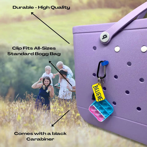A close-up of a purple bag features the Chatelet Manufacturing BOGLETS Carabiner Keys Holder Charm Accessory with a BELIEVE pop-it keychain. In the background, a woman and three kids stand in a grassy field. Text highlights this fun Bogg Bag accessory’s features.