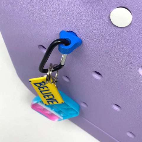 The Chatelet Manufacturing BOGLETS Bogg Bag Carabiner Keys Holder Charm Accessory in blue, with a keyring, yellow BELIEVE tag, and blue-pink pop-it toy, adds fun and secure organization to your Bogg Bag on a lavender pegboard.
