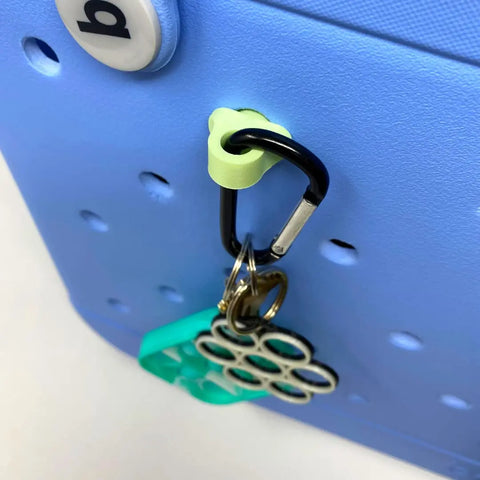 The Chatelet Manufacturing BOGLETS Carabiner Keys Holder Charm Accessory in black secures your keys and teal keychain to your Bogg Bag with style, keeping valuables organized on the blue perforated bag surface.