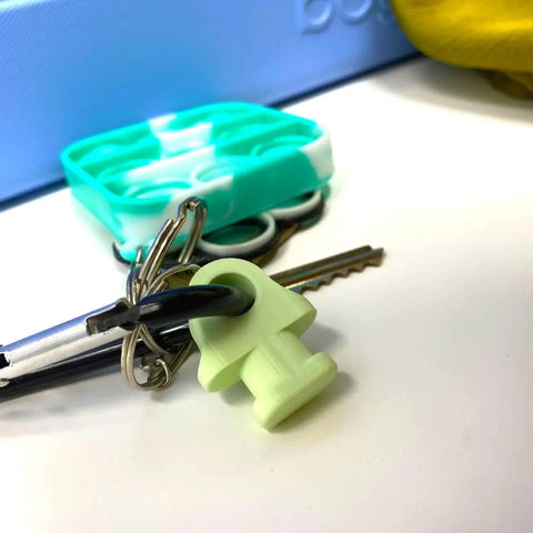 The BOGLETS by Chatelet Manufacturing, a green mushroom-shaped carabiner keys holder charm, rests near a blue and white silicone fidget toy on a white surface with yellow fabric in the background.