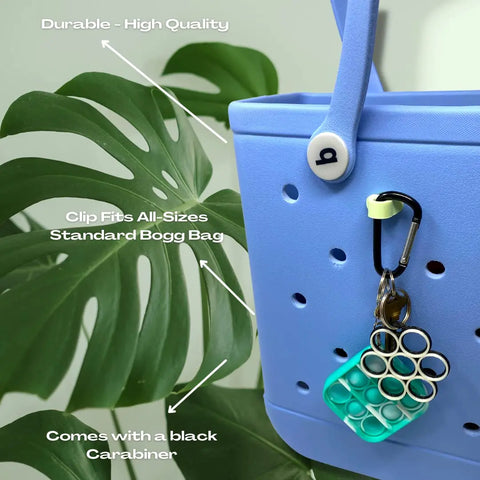 Close-up of a blue bag with a Chatelet Manufacturing BOGLETS Carabiner Keys Holder Charm Accessory, shown securely holding a green and blue bubble pop toy. Text highlights its durability, Bogg Bag compatibility, and the included carabiner.