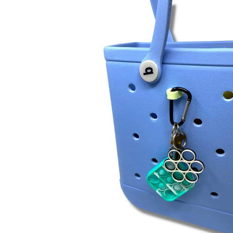 A light blue tote bag with a green and white pop-it keychain attached by a black BOGLETS Bogg Bag Carabiner Keys Holder Charm Accessory from Chatelet Manufacturing, keeping your valuables secure and organized in style.