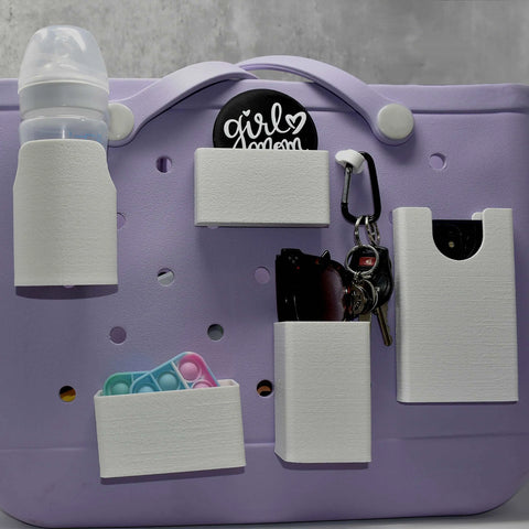 A lavender tote bag with white Chatelet Manufacturing BAGLETS accessories, including a phone case holder, key & wallet holder, carabiner key holder, bottle holder accessory, and sunglasses holder—plus a pop-it toy and a girl mom pin.